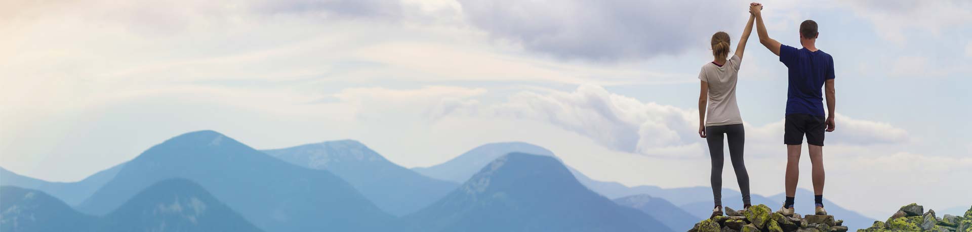 Back view of young tourist couple, athletic man and slim girl standing with raised arms holding hands on rocky mountain